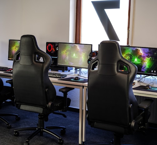 A row of gaming stations equipped with large monitors, gaming chairs, keyboards, and headphones. The setup is organized and ready for gaming, featuring a colorful screen display. The room has a modern and tech-savvy ambiance.