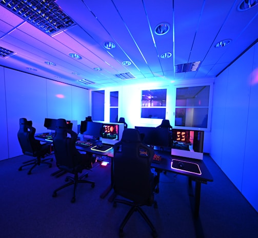 A modern gaming setup with multiple high-backed gaming chairs and monitors arranged in a room with a cool blue lighting. The computers have illuminated keyboards and glowing screens, creating a high-tech ambiance.