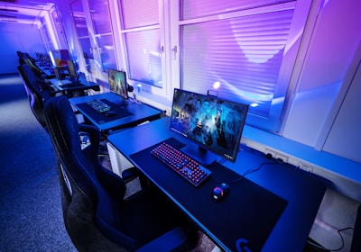 A row of modern gaming setups is arranged neatly along a window-lined wall. Each station includes a high-resolution monitor displaying vivid graphics, a mechanical keyboard with colored lighting, a gaming mouse, and an ergonomic chair. The room is illuminated with ambient blue and purple lights, creating a sleek and futuristic atmosphere.