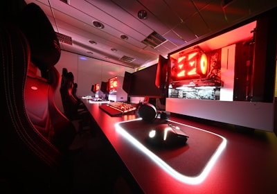A modern gaming setup featuring several high-end computers with illuminated red interior lighting. The room is equipped with gaming chairs exhibiting a sleek black and red design. Multiple monitors are arranged on a long desk, and peripheral devices like keyboards and mice are uniformly placed, reflecting an ambiance ideal for gaming or competitive play.