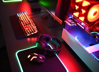 A gaming setup with a mechanical keyboard featuring multicolor RGB lighting, a glowing mouse on a mousepad with illuminated edges, a set of headphones, and a high-end gaming PC with a transparent side panel showcasing internal components and red LED lights.