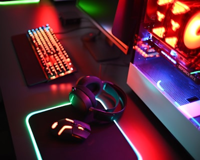 A gaming setup with a mechanical keyboard featuring multicolor RGB lighting, a glowing mouse on a mousepad with illuminated edges, a set of headphones, and a high-end gaming PC with a transparent side panel showcasing internal components and red LED lights.
