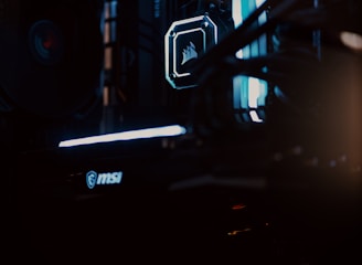 A close-up view of a computer gaming rig illuminated with blue LED lights. The interior shows components like cooling fans at the top, a glowing logo on a central component, and an MSI branded part at the bottom. The scene is dark, emphasizing the glow of the lights.