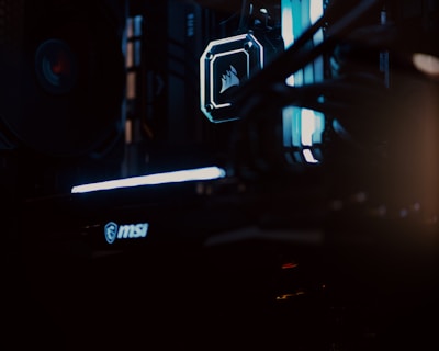 A close-up view of a computer gaming rig illuminated with blue LED lights. The interior shows components like cooling fans at the top, a glowing logo on a central component, and an MSI branded part at the bottom. The scene is dark, emphasizing the glow of the lights.