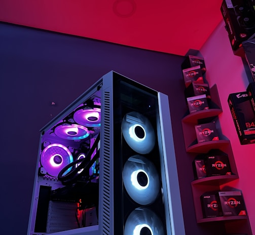A gaming computer setup featuring a high-performance PC tower with multiple RGB fans lit in blue and purple hues. The case has a modern design with transparent panels showcasing internal components. In the background, shelves are stocked with various Ryzen and other gaming components under a vibrant red and blue ambient light.