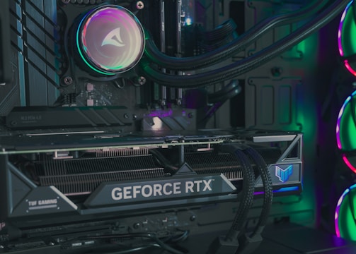 The image depicts the inside of a computer case featuring a high-performance graphics card labeled 'GEFORCE RTX' with RGB lighting. The cooling fans are illuminated with vibrant colors, creating a visually striking look. Various cables and components are neatly arranged, highlighting advanced hardware and a focus on aesthetics.